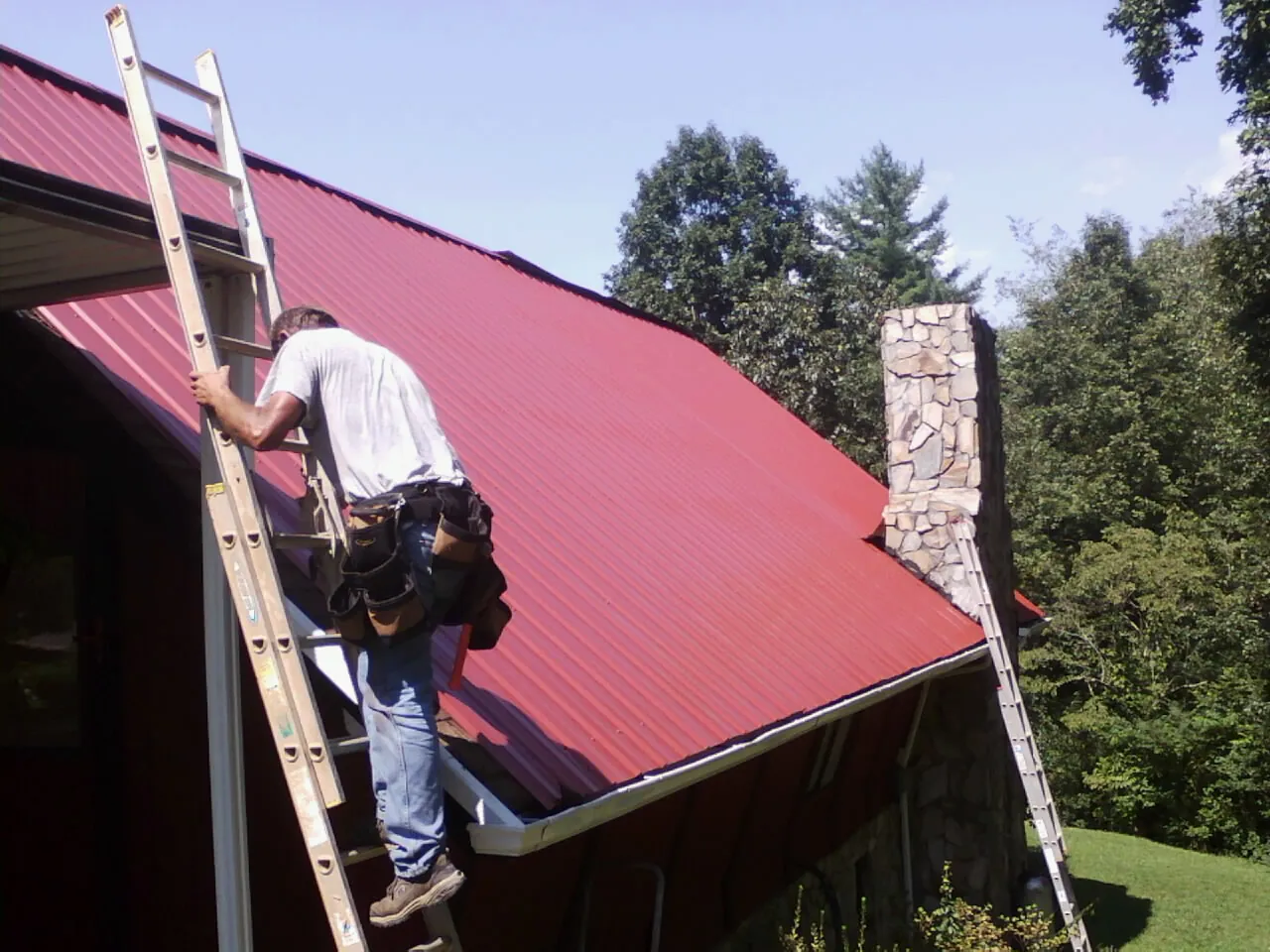 Roofer on a ladder beside a red metal roof and stone chimney — Soffit & Fascia Repair by Roofing Brothers in East Brookfield